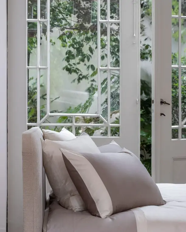 Satijnen kussens in taupe en ivoor op een linnen bed, bij tuindeuren met glas in lood
