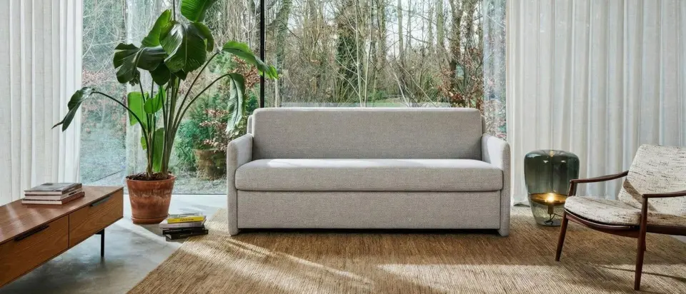 De slaapbank Dexter in een glazen woonkamer met uitzicht op het bos, met een grote plant en een houten dressoir