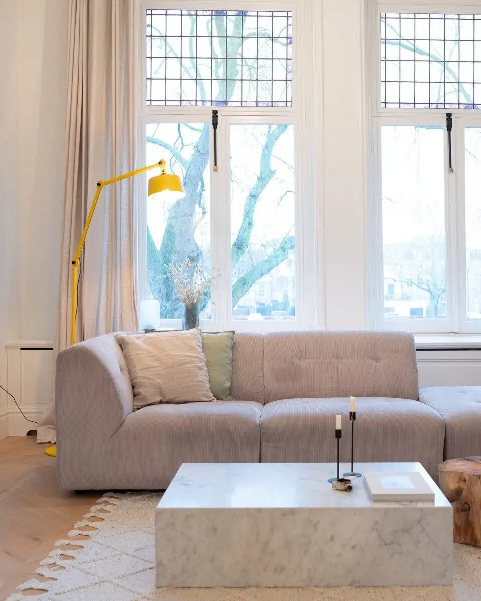 Een gele vloerlamp met twee scharnierende armen in een lichte woonkamer met hoge ramen