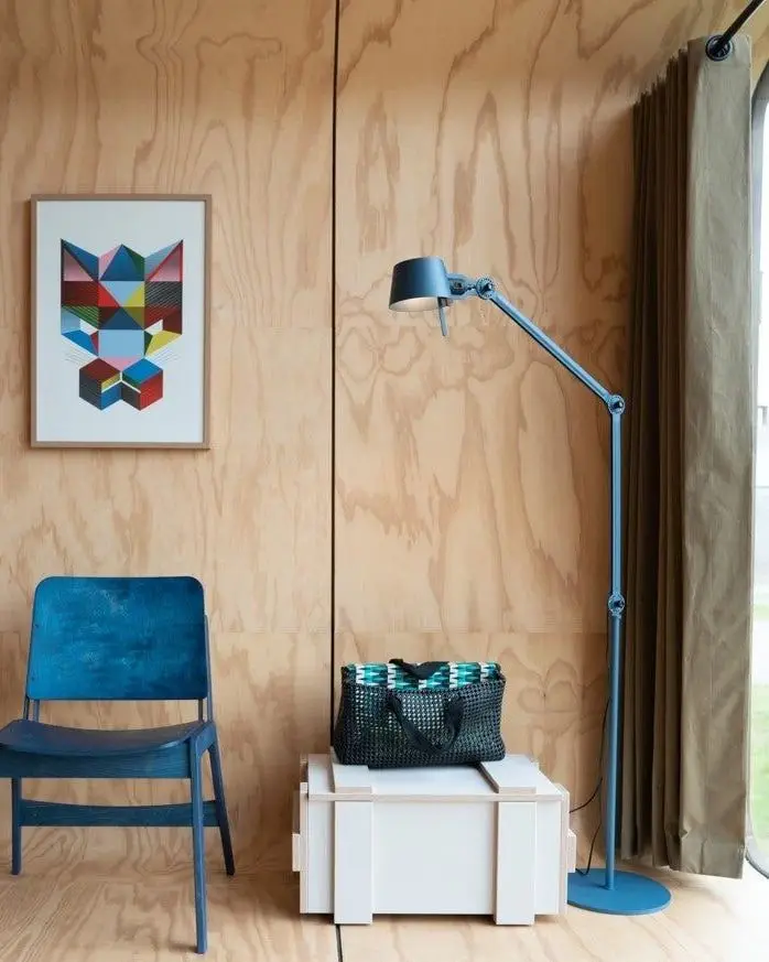 Een blauwe vloerlamp tegen een wand van multiplex, naast een blauwe stoel
