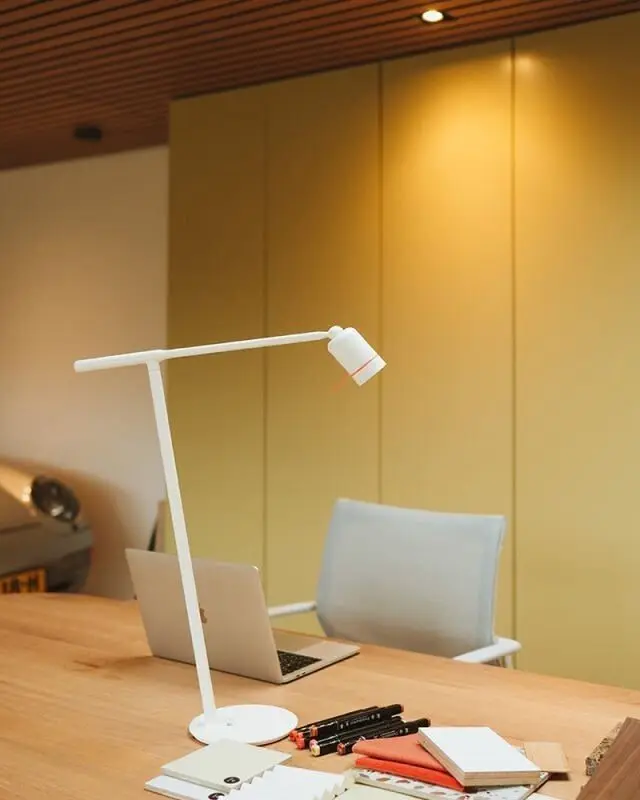 Een witte bureaulamp op een houten werkblad voor een gele wand