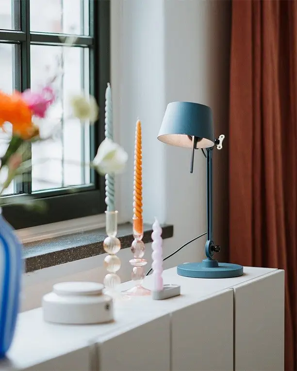 Een blauwe bureaulamp op een witte kast bij het raam, naast een vaas met bloemen