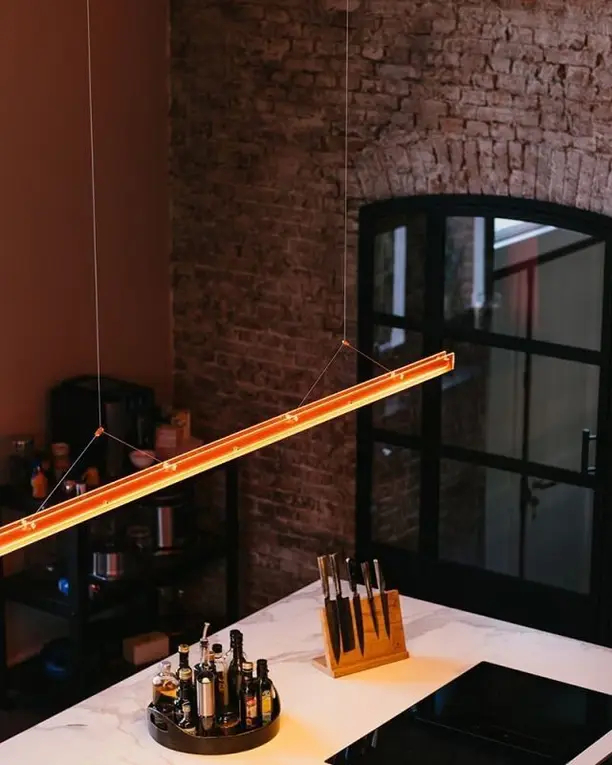 Een lange, dunne lamp met een warme lichtlijn boven een marmeren keukeneiland voor een bakstenen boog