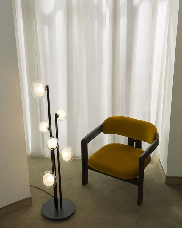 Een staande lamp met glazen druppels naast een okergele stoel voor lichte vitrage