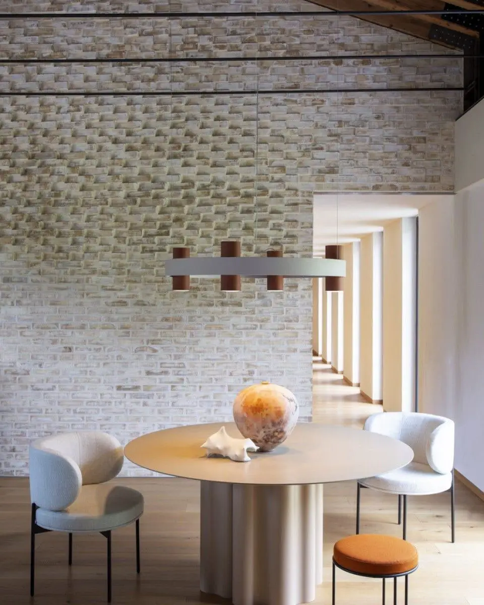 Een ronde hanglamp met terracotta cilinders boven een ronde tafel met bouclé stoelen, voor een lichte bakstenen wand