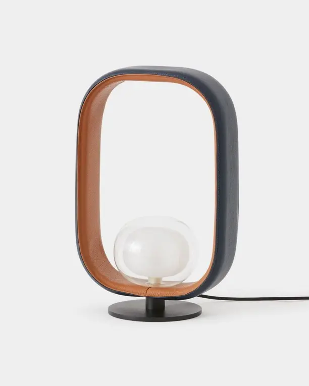Een tafellamp met een ovale donkere omlijsting met een koperkleurige binnenkant rond een glazen bol, op een lichte achtergrond