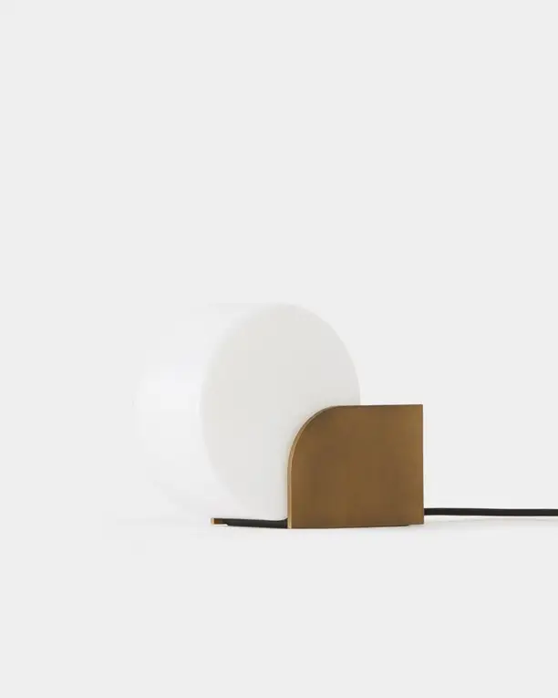 Een tafellamp met een witte glazen bol voor een gebogen bronskleurig scherm, op een lichte achtergrond