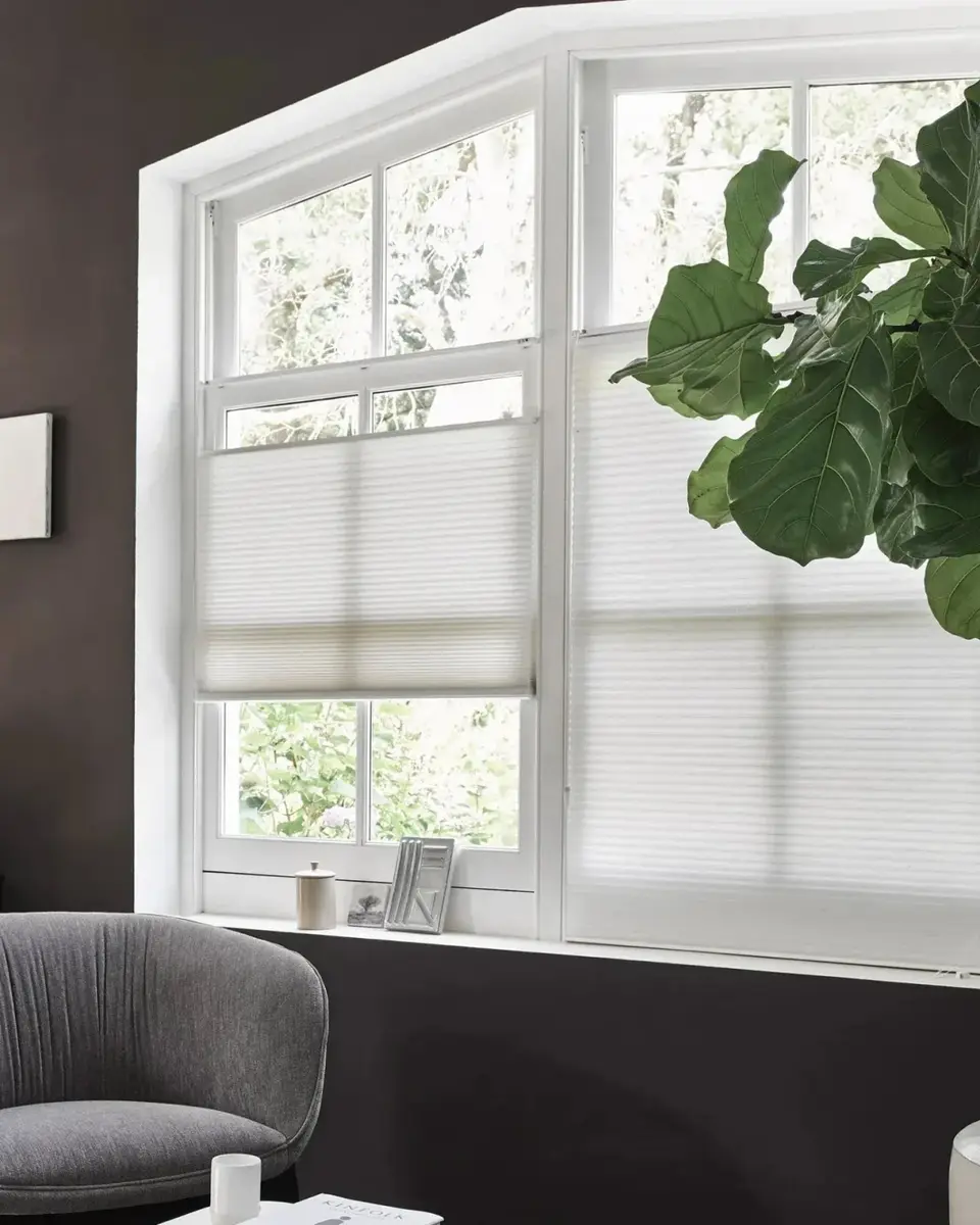 Lichtdoorlatende plissé in een raam met een schuine bovenkant, voor een donkerbruine wand met een grijze fauteuil