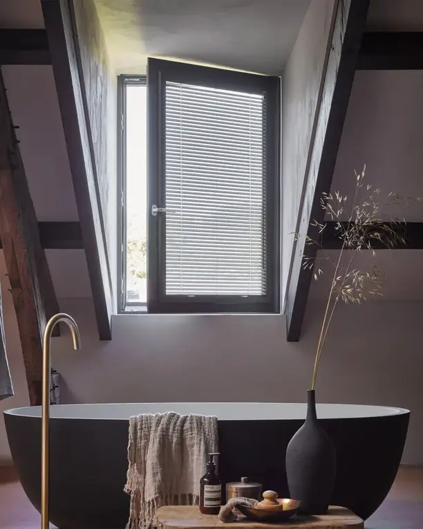 Zolderbadkamer met een vrijstaand bad en een aluminium jaloezie in het raam tussen de balken