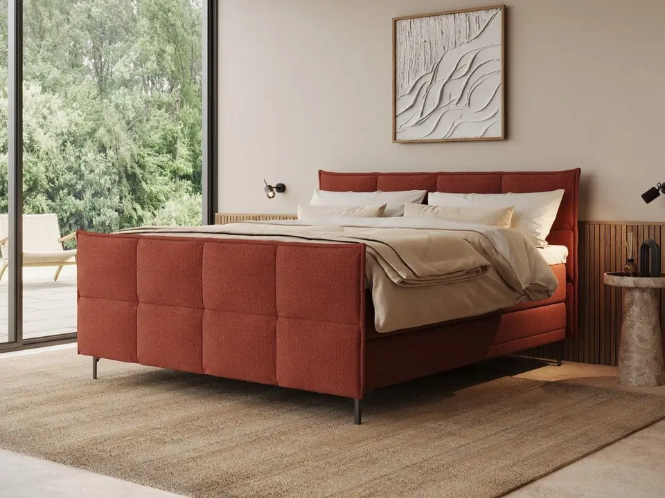 Een boxspring in terracotta stof met een geruit hoofdbord en voeteneinde, bij een groot raam met bomen