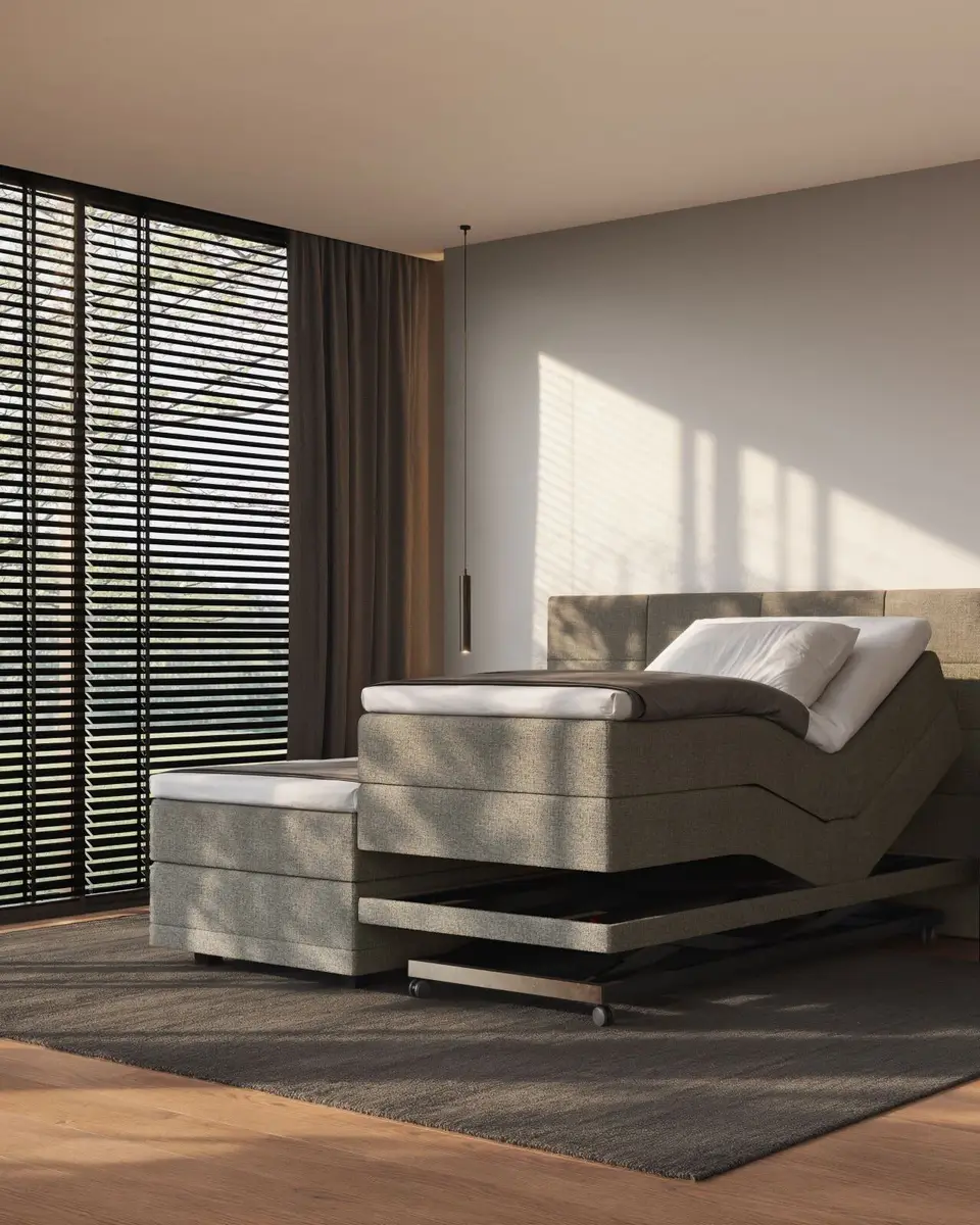 Een elektrisch verstelbare boxspring in taupe stof met het hoofdeinde omhoog op een hoog-laagframe, bij een raam met jaloezieën