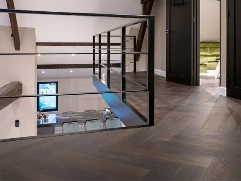 Donkere visgraatvloer langs een vide met een glazen balustrade en houten balken