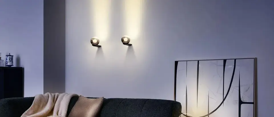 Twee Luna-wandlampen werpen warm licht op een grijze wand boven een donkere bank met een lichte plaid