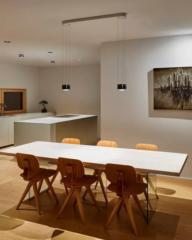 Twee zwarte hanglampen boven een lange witte eettafel met houten stoelen, met een schilderij aan de wand