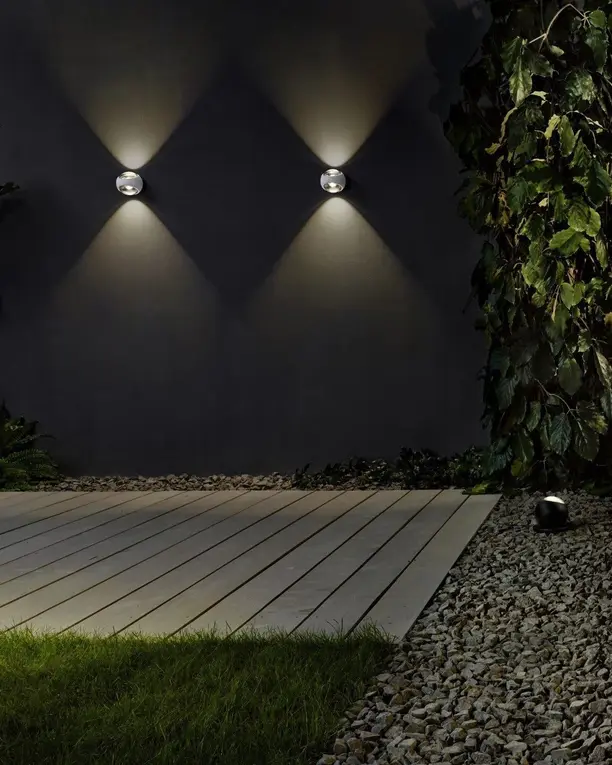 Twee wandlampen werpen licht naar boven en naar beneden op een donkere tuinmuur naast een terras