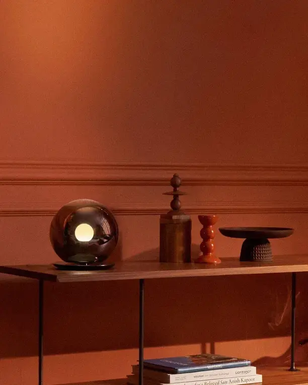 Een glazen Luna-tafellamp op een smalle console voor een warme terracotta wand, bij avond
