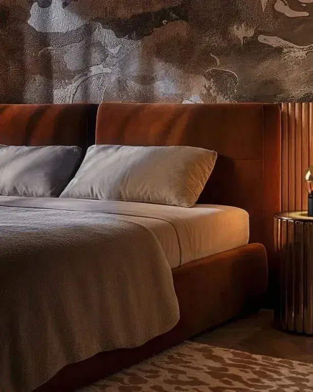 Een Luna-tafellamp op een bronzen nachtkastje naast een bed met een roestkleurig hoofdbord