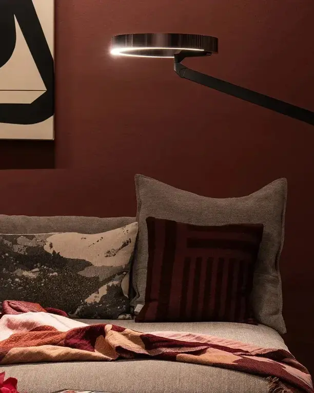 Een Mito-leeslamp buigt over een bank met kussens en een plaid, voor een ossenbloedrode wand