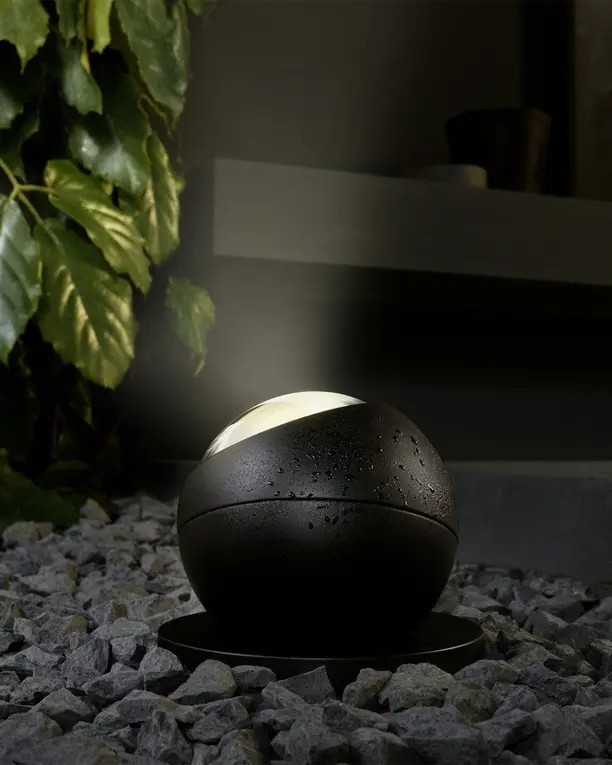 Een donkere bollamp tussen grind en grote bladeren in een tuin bij avond