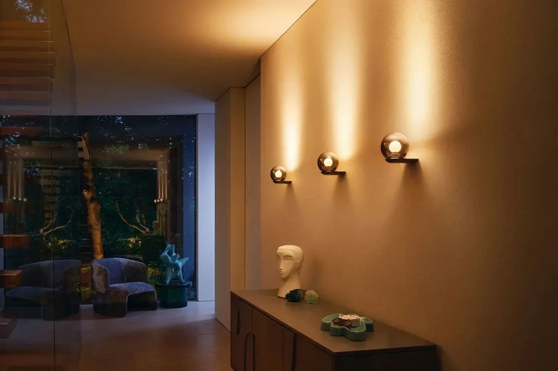 Drie ronde wandlampen verlichten een warme wand boven een notenhouten dressoir, met zicht op de tuin