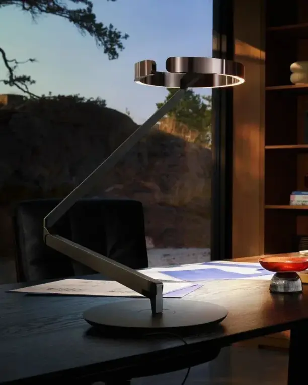 Een bureaulamp met een ronde lampkop op een lange arm, op een bureau bij een raam in de schemer