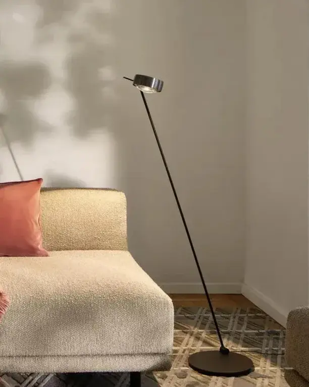 Een slanke leeslamp naast een lichte bank met kussens, voor een wand met een groot schilderij