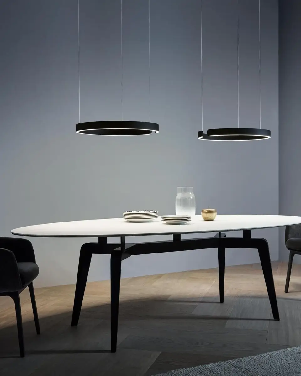Twee dunne lichtringen van Occhio Mito boven een ovale witte eettafel met zwarte stoelen, tegen een grijze wand