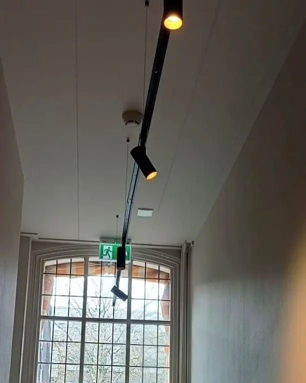 Een lange witte gang met spots aan een zwarte rail en een raam aan het eind