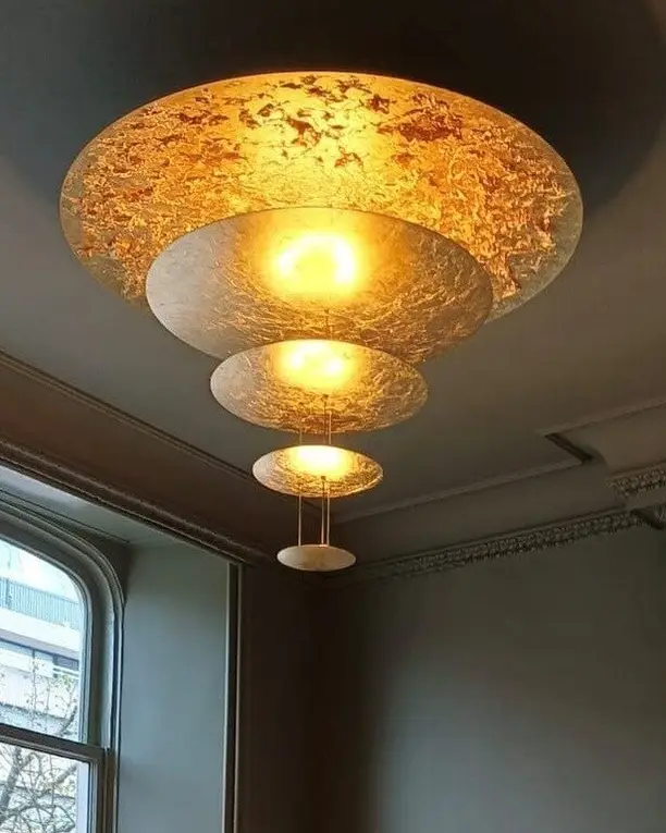 Een getrapte hanglamp met bladgoud brandt in een groengrijze kamer met hoge ramen en een kast