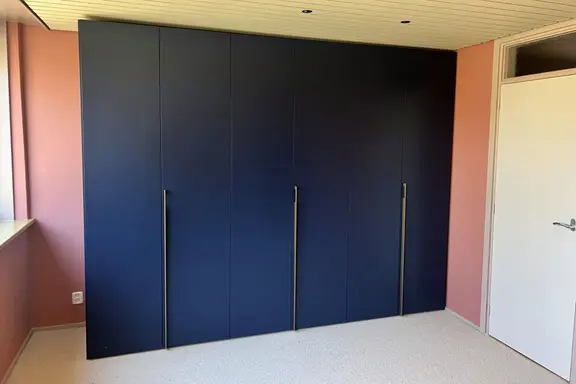 Een matte donkerblauwe kledingkast op maat van vloer tot plafond, met lange greeplijsten, tussen twee roze wanden