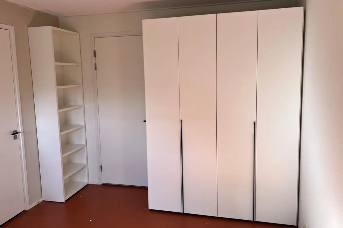 Een witte kledingkast met vier deuren en een open boekenkast in een slaapkamer met een terracotta vloer