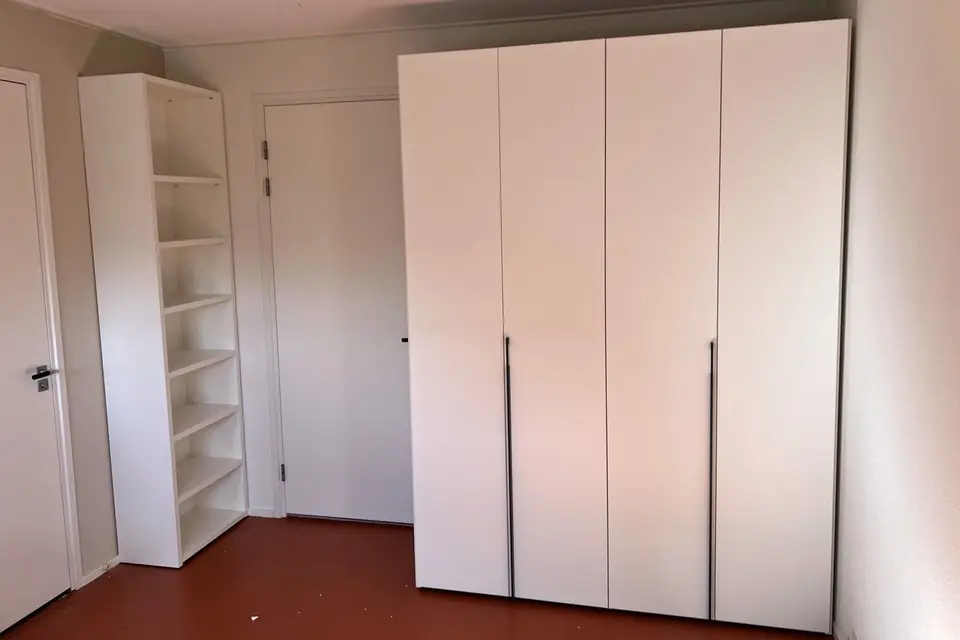 Een witte kledingkast met vier deuren en een open boekenkast in een slaapkamer met een terracotta vloer