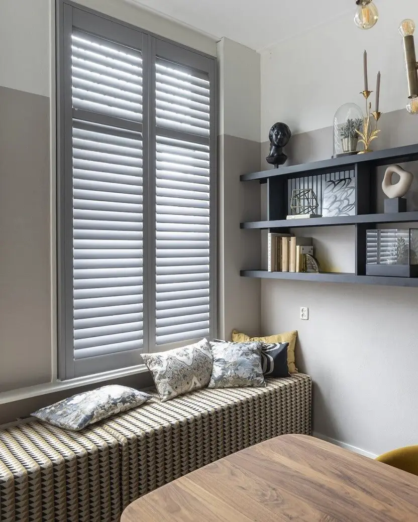 Grijze shutters voor een hoog raam, met een zitbank in de vensterbank en open wandplanken