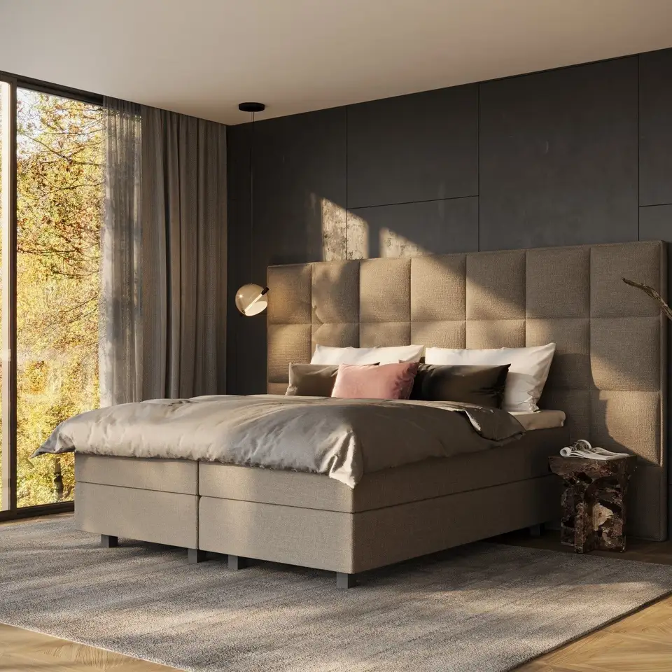 Boxspring met een hoog gestoffeerd hoofdbord tegen een donkere wand, laag zonlicht door een raam met herfstbomen