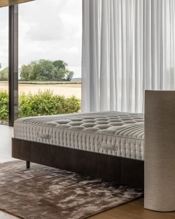 Een getufte matras op een geribbeld notenhouten onderstel in een glazen paviljoen met uitzicht op het land