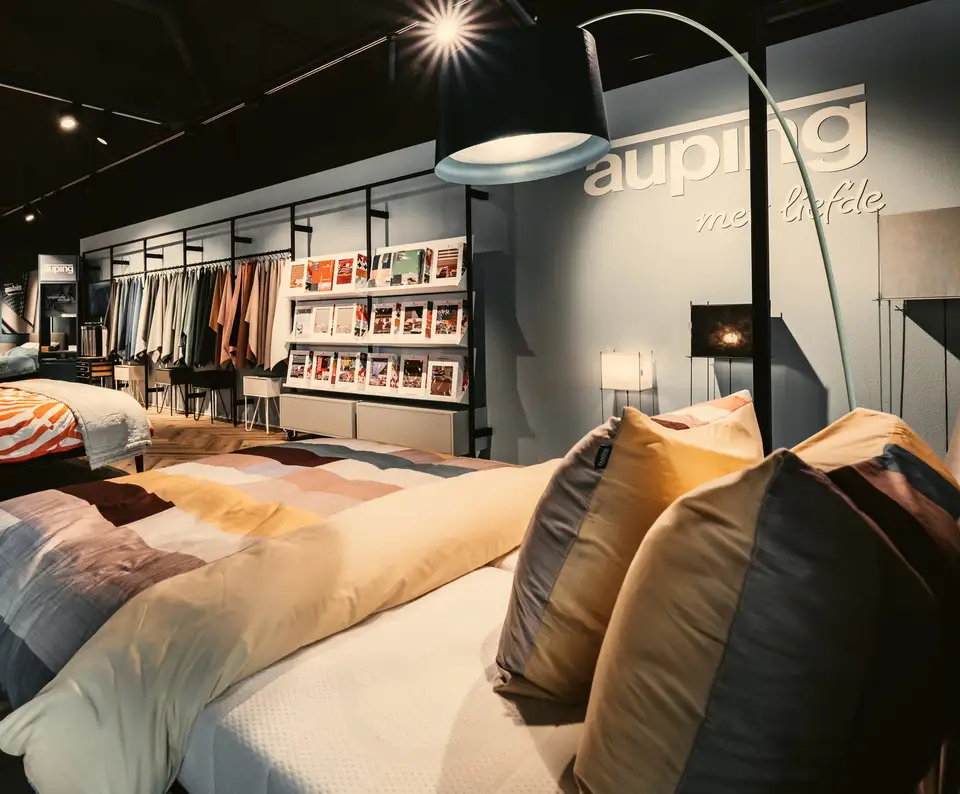 Onze Auping Studio: een opgemaakt bed met kleurige kussens onder een booglamp, voor een blauwgrijze wand met het Auping-logo en een rek met bedtextiel