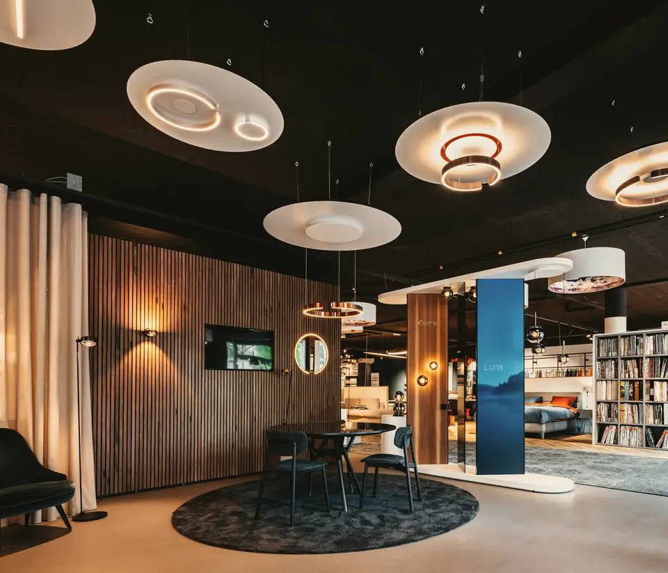 Onze showroom aan het Zuiderplein: witte schijven met lichtringen boven een ronde tafel, een houten lamellenwand en de Occhio-zuil, met daarachter bedden en een wand vol beddengoed