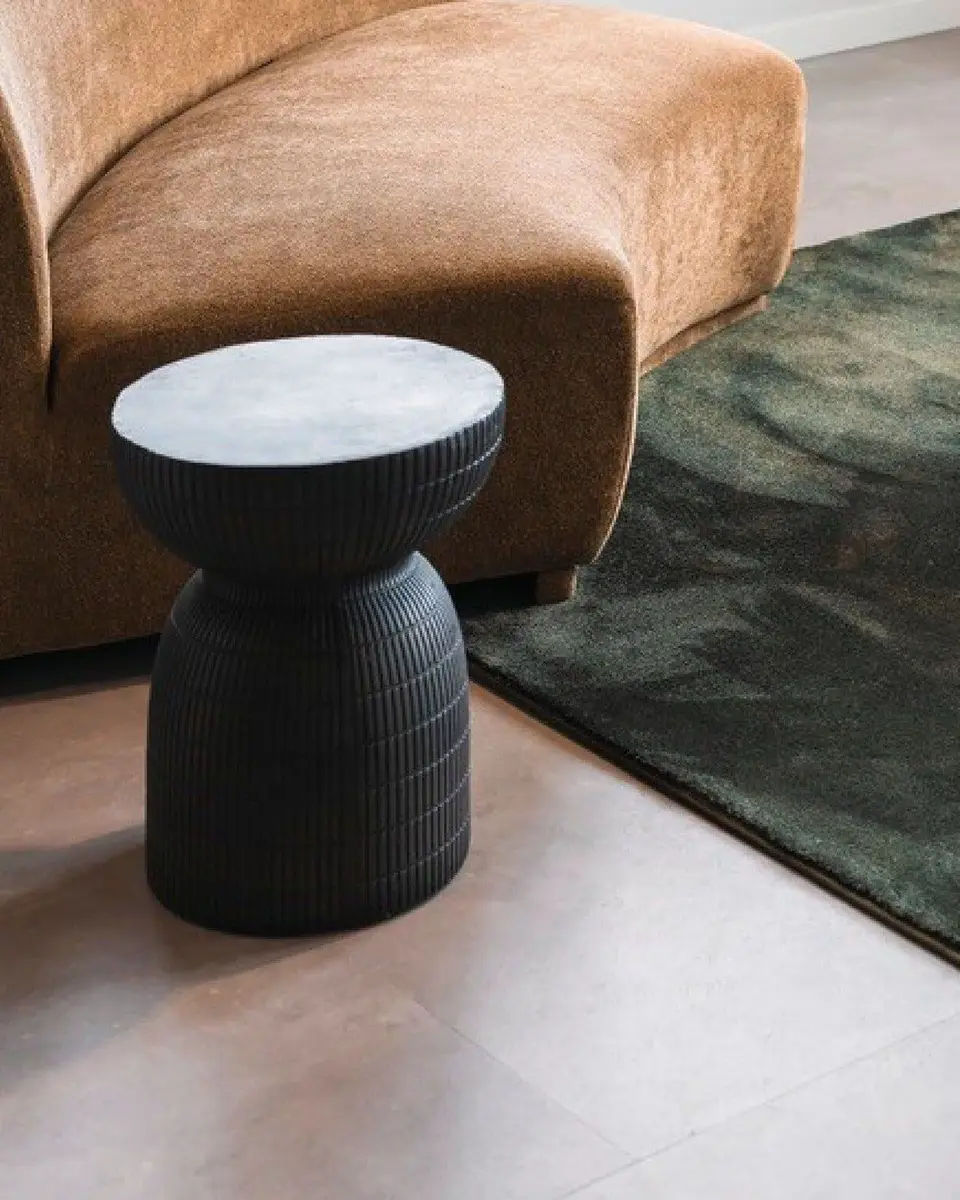Cognackleurige fauteuil, een zwarte kruk en een groen karpet op grote pvc-tegels in betonlook